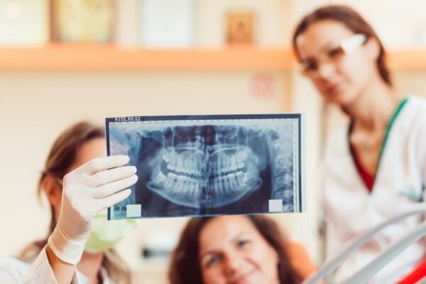 Two dentists wearing protective gear, and a patient examining an x-ray of the patient's teeth in a dental office.