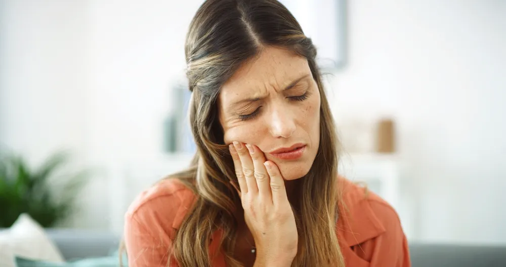 A woman sitting on a couch, holding her jaw in discomfort, indicating a toothache.